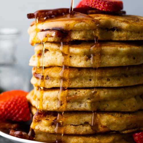 side view of a large stack of bacon pancakes on a plate with syrup being drizzled over and fresh strawberries