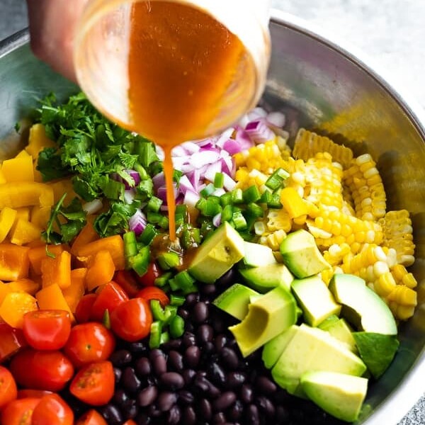 dressing being poured out of a mason jar over black bean and corn salad in large metal bowl
